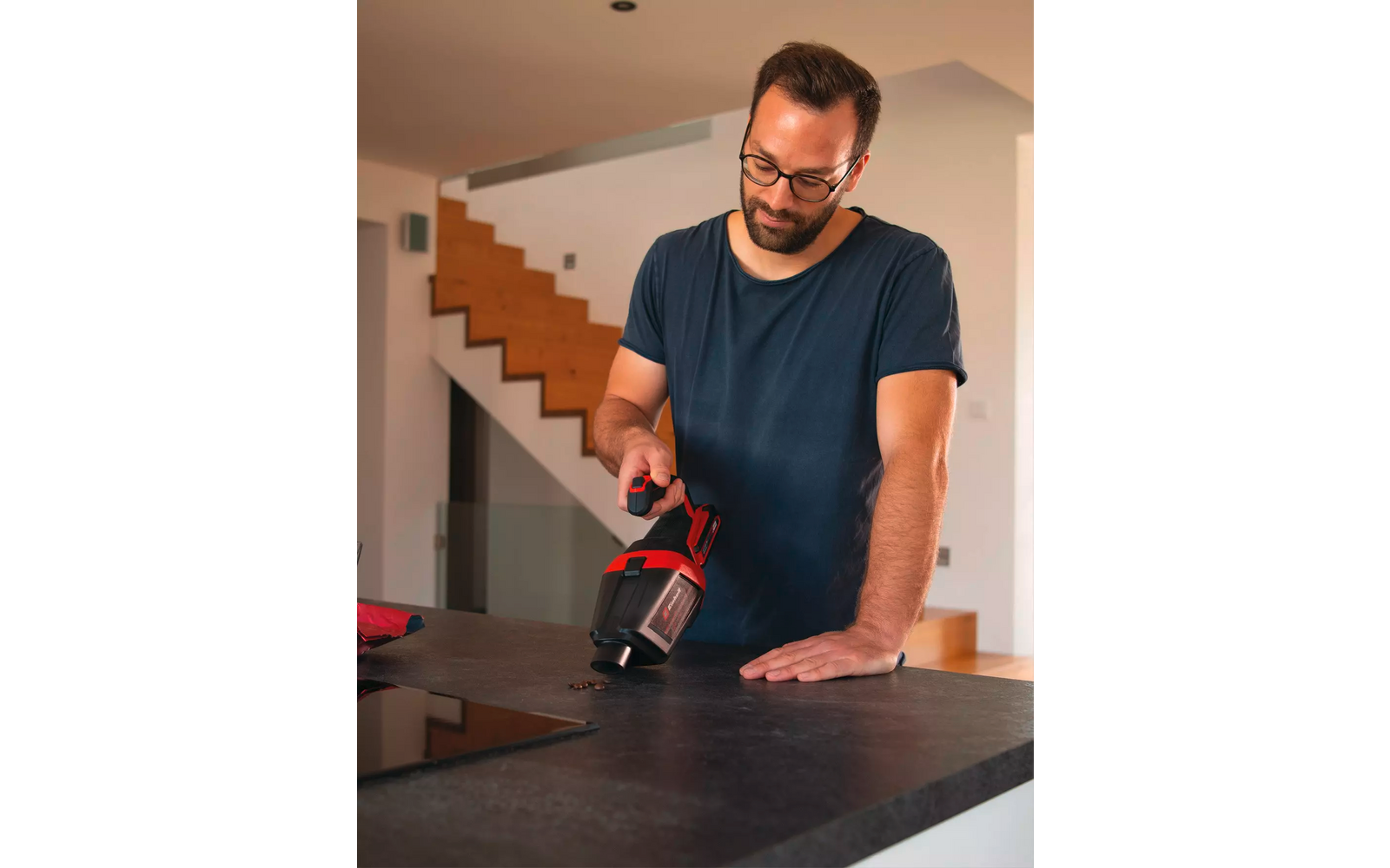 Mann mit Brille und Bart hält roten und schwarzen Handsauger über dunkler Küchenarbeitsplatte, Treppe im Hintergrund