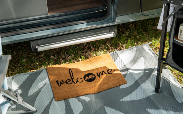 Paillasson avec le mot 'welcome' devant la porte ouverte d'un camping-car, sur un tapis d'extérieur à motif et de l'herbe à côté