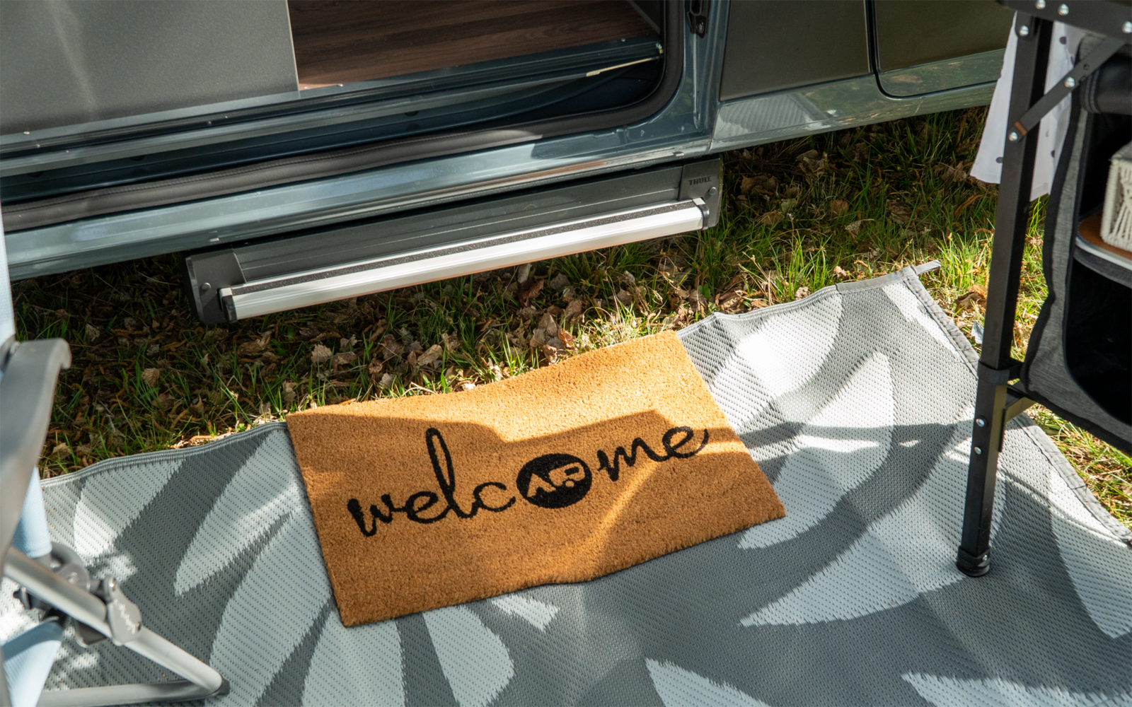 Paillasson avec le mot 'welcome' devant la porte ouverte d'un camping-car, sur un tapis d'extérieur à motif et de l'herbe à côté
