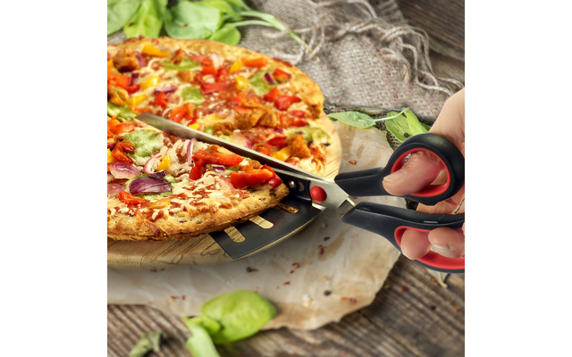 Main tenant des ciseaux noirs à manche rouge coupant une pizza garnie de fromage, tomates, oignon rouge, poivron et basilic sur une planche en bois