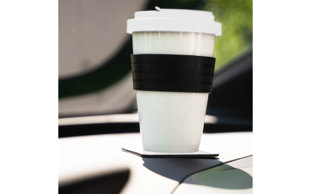 Weißer Becher mit schwarzem Silikonband und weißem Deckel auf einem quadratischen Untersetzer im Getränkehalter des Armaturenbretts, unscharfer Außenhintergrund