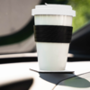 Weißer Becher mit schwarzem Silikonband und weißem Deckel auf einem quadratischen Untersetzer im Getränkehalter des Armaturenbretts, unscharfer Außenhintergrund