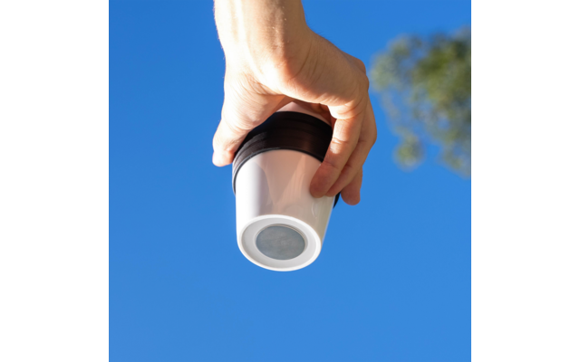 Hand hält weißen Becher mit schwarzem Deckel vor blauem Himmel, rechts unscharfer Baum