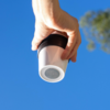 Hand hält weißen Becher mit schwarzem Deckel vor blauem Himmel, rechts unscharfer Baum