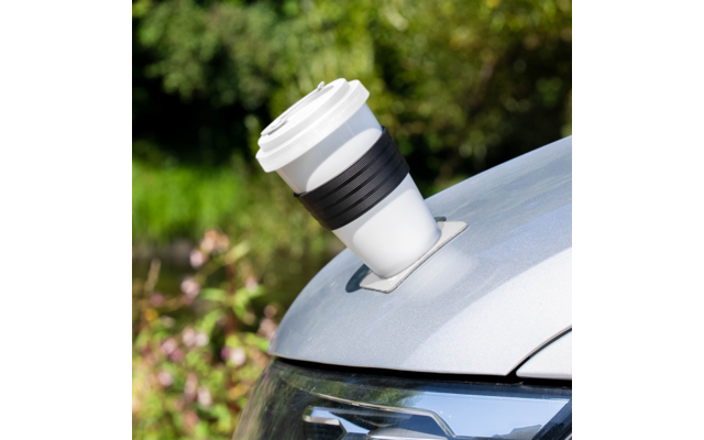 Weißer Becher mit Deckel und schwarzem Silikonband in einem eingelassenen Getränkehalter auf der silbernen Motorhaube eines Autos, unscharfer grüner Hintergrund