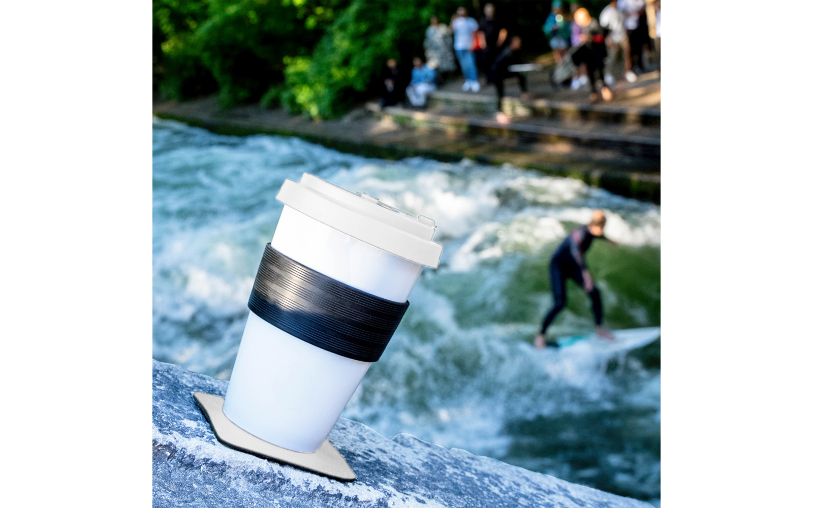 Pappbecher mit Kunststoffdeckel und schwarzem Halteband auf einem Felsen am Ufer eines Flusses; im Hintergrund eine Person auf einem Surfbrett und mehrere Personen an der Flussbank