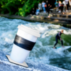 Pappbecher mit Kunststoffdeckel und schwarzem Halteband auf einem Felsen am Ufer eines Flusses; im Hintergrund eine Person auf einem Surfbrett und mehrere Personen an der Flussbank