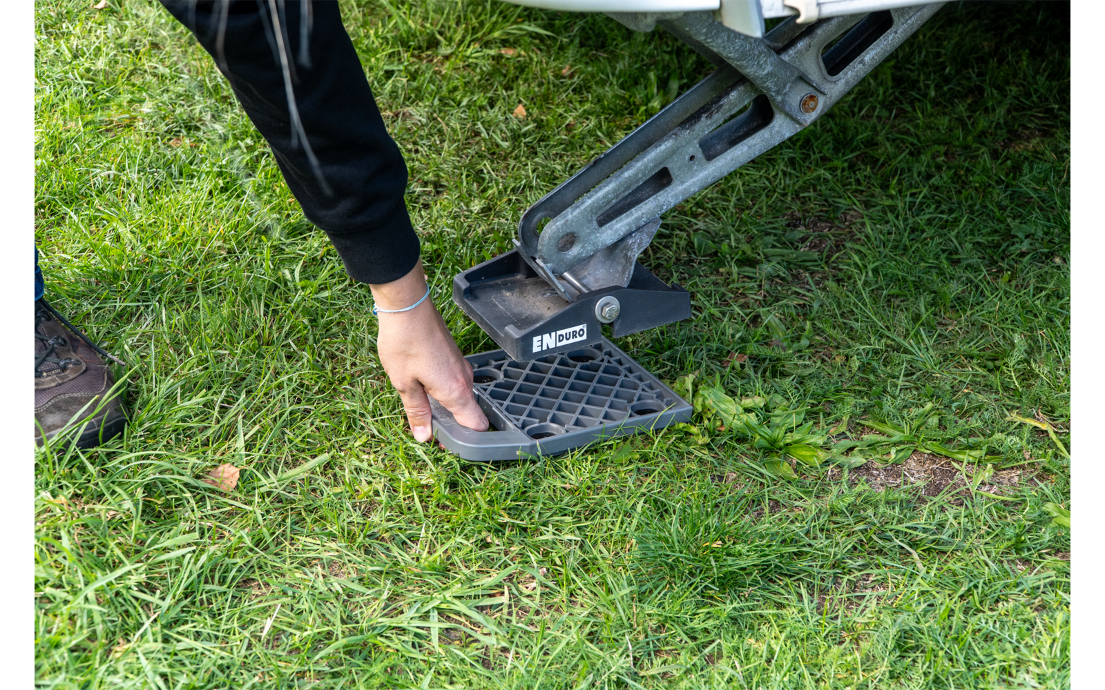Hand setzt ein quadratisches Kunststoffgitter unter eine Metallstütze eines Fahrzeugs auf Gras