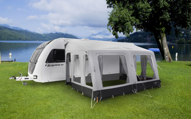 Wohnwagen mit angebautem weiß-grauem Vorzelt auf einer grünen Wiese, im Hintergrund ein See und Berge unter blauem Himmel mit Wolken