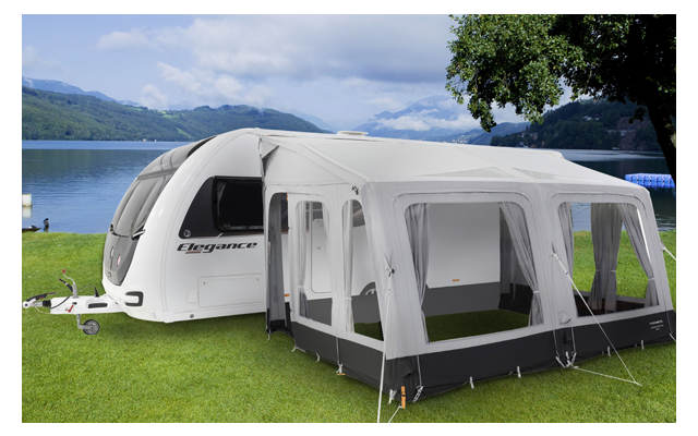 Wohnwagen mit angebautem weiß-grauem Vorzelt auf einer grünen Wiese, im Hintergrund ein See und Berge unter blauem Himmel mit Wolken