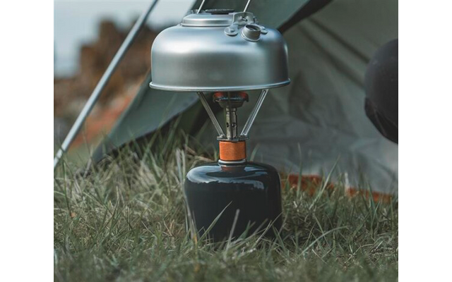 Kleiner Gaskocher auf schwarzer Gaskartusche im Gras vor einem Zelt, silberner Topf auf dem Brenner