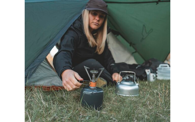 Person sitzt in einem geöffneten Zelt mit der Hand an einem Campingkocher; neben der Person ein Wasserkessel und Gepäck auf Gras