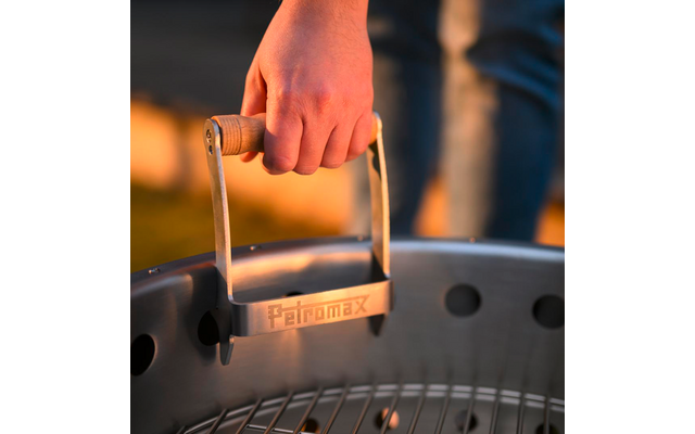 Hand hält einen hölzernen Griff an einem runden Metallgrill; Metallplatte mit Schriftzug 'Petromax'; unscharfe Beine in Jeans im Hintergrund