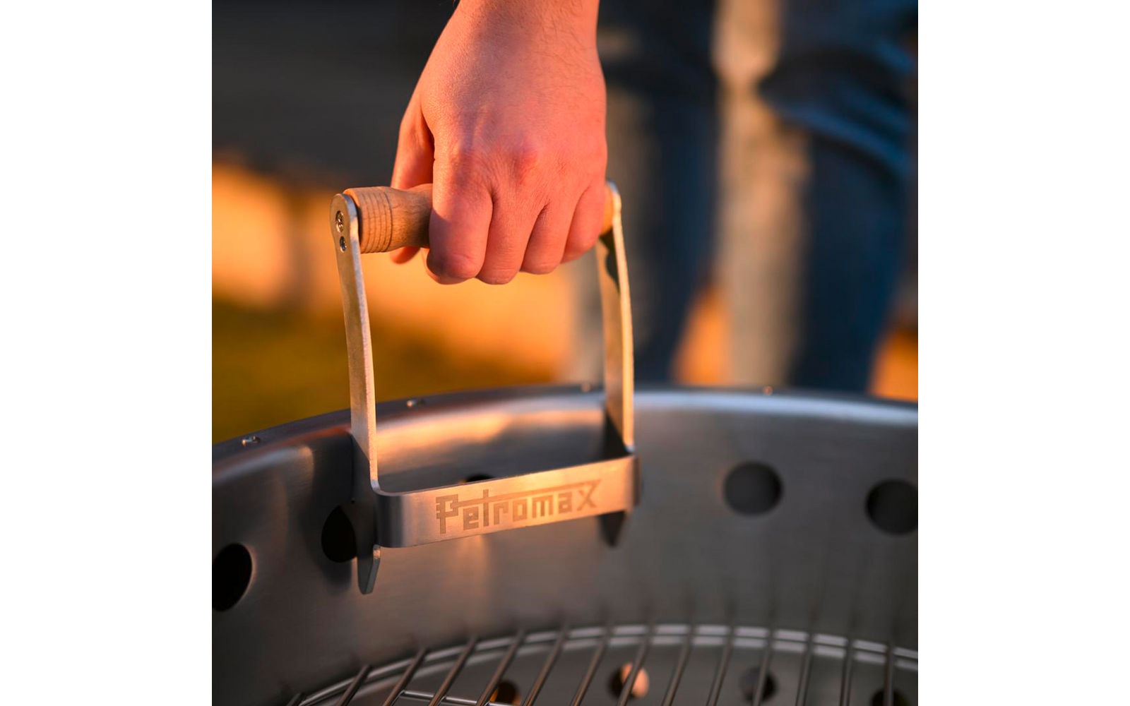 Hand hält einen hölzernen Griff an einem runden Metallgrill; Metallplatte mit Schriftzug 'Petromax'; unscharfe Beine in Jeans im Hintergrund