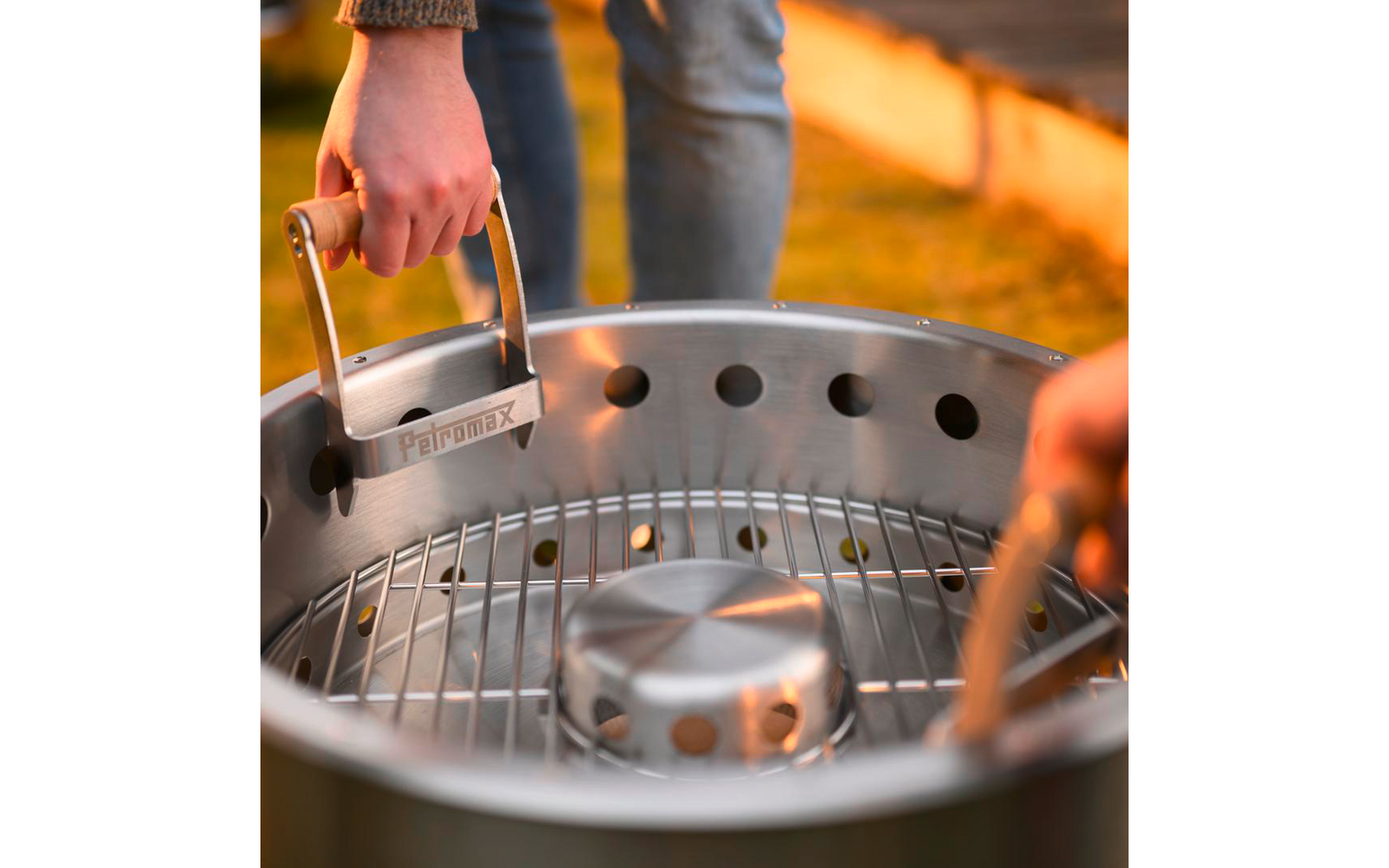 Zwei Hände über einem runden Edelstahlgrill; eine Hand hält einen rechteckigen Metallgriff, die andere eine Grillzange; sichtbarer Grillrost, unscharfer Hintergrund mit Gras und warmem Licht