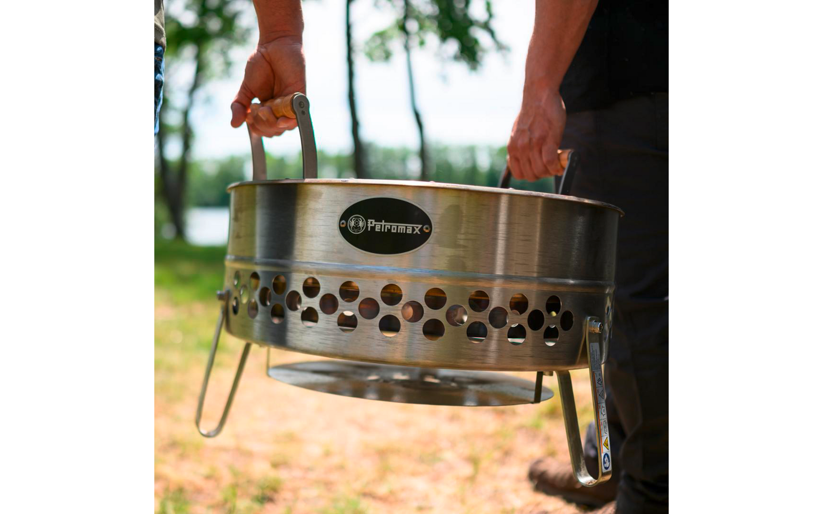 Zwei Personen, nur Hände und Unterarme sichtbar, tragen im Freien einen runden Edelstahlgrill mit Griffen und der Aufschrift Petromax