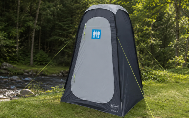 Tenda grigia alta e rettangolare con tetto arrotondato e porta con cerniera; cartello blu sulla porta con pittogrammi maschile e femminile; tiranti gialli; su erba vicino a un ruscello, circondata da alberi