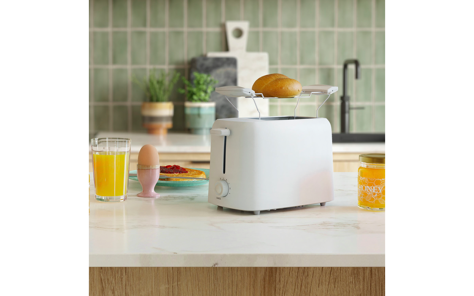 Weißer Toaster auf Küchenarbeitsplatte mit Brötchen im hochgeklappten Rost; Glas Orangensaft, Ei im rosa Eierbecher, kleines Tellerchen mit roter Marmelade, Honigglas, Topfpflanzen und grüne Kachelrückwand