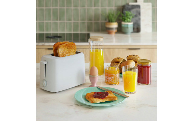 Toaster mit zwei gerösteten Brotscheiben, Teller mit Toast bestrichen mit roter Marmelade und Messer, weichgekochtes Ei im Eierbecher, Glas und Karaffe mit Orangensaft, zwei Marmeladengläser, Brötchen auf der Arbeitsplatte, grüne Fliesenrückwand