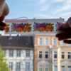 Zwei Hände halten ein transparentes Band mit orangenen und roten Blüten vor einem Häuserhintergrund