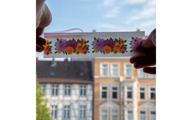 Zwei Hände halten ein transparentes Band mit orangenen und roten Blüten vor einem Häuserhintergrund