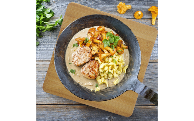 Gusseiserne Pfanne auf Holzbrett mit cremiger Sauce, zwei gebräunte Medaillons, Spätzle, Pfifferlingen und Petersilienblättern