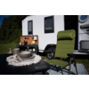 Caravane blanche sur l'herbe avec porte ouverte, chaise de camping verte pliable au premier plan, table pliante avec assiettes et bol sur un napperon à franges, cuisine de camping mobile avec ustensiles et cafetière, tapis d'extérieur à motifs, arbres en arrière-plan