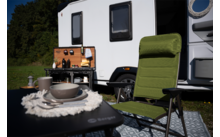 Weißer Wohnwagen auf einer Wiese mit offenem Türbereich, grüner Campingstuhl im Vordergrund, Klapptisch mit Tellern und Schüssel auf einem Tischläufer mit Fransen, mobile Campingküche mit Utensilien und Kaffeemaschine, gemusterter Außenteppich, Bäume