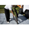 Persoon vouwt een opgevouwen groene campingstoel uit op een patroonmat naast een camper; handen, benen en witte schoenen zichtbaar