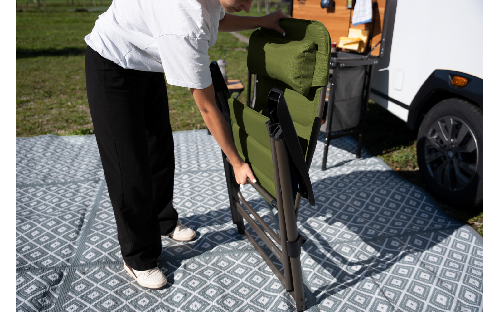 Persoon vouwt een opgevouwen groene campingstoel uit op een patroonmat naast een camper; handen, benen en witte schoenen zichtbaar