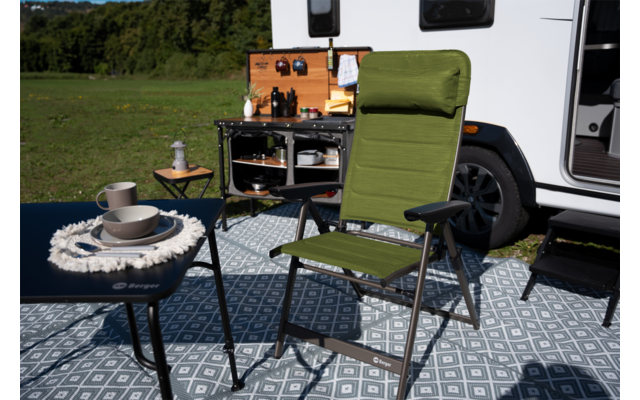 Groene opvouwbare campingstoel voor een witte camper, kleine tafels met kopjes en kommen, open campingkeuken aan de camper, patroonmat op de grond en grasveld op de achtergrond