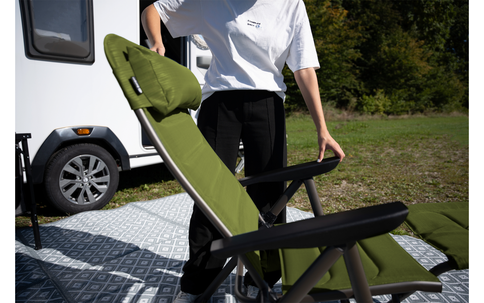 Bovenlichaam van een persoon zonder zichtbaar gezicht die een olijfgroene campingstoel met zwart frame vasthoudt op een patroonmat naast een witte caravan; gras en bomen op de achtergrond