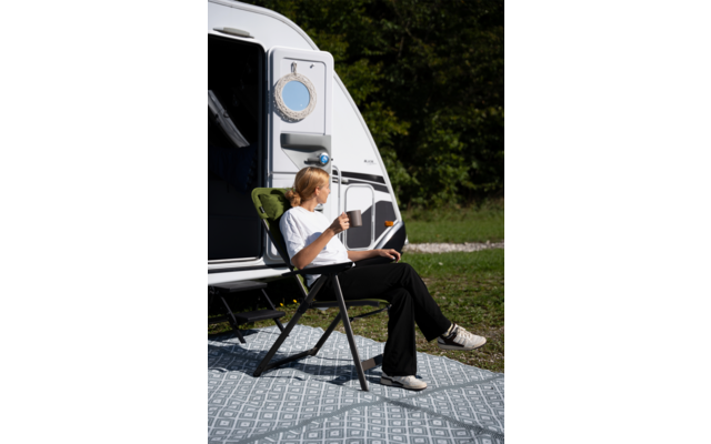 Persoon zit in een opvouwbare campingstoel voor een open caravandeur, houdt een mok en een tablet op schoot; patroonmat op het gras, bomen op de achtergrond