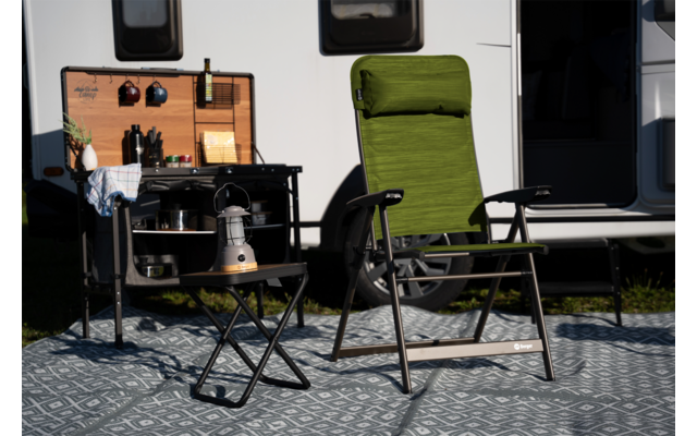 Groene opvouwbare relaxstoel voor een witte caravan; patroonmat op de grond; klein zwart opvouwbaar bijzettafeltje met metalen ketel; daarnaast een opvouwbare keukenunit met houten paneel, flessen, bestek en pannen