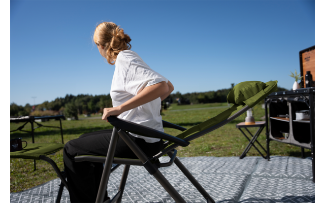 Persoon met blonde knot, wit T‑shirt en zwarte broek zit op een opvouwbare stoel op een gepatineerde/gevlekte ondergrondmat en kijkt naar rechts; op de achtergrond weiland en blauwe lucht