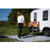 Personne plie une chaise de camping verte à côté d'une caravane blanche avec un compartiment extérieur ouvert, sur un tapis à motifs, herbe et arbres en arrière-plan