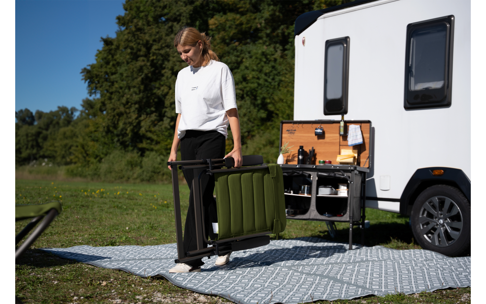 Personne plie une chaise de camping verte à côté d'une caravane blanche avec un compartiment extérieur ouvert, sur un tapis à motifs, herbe et arbres en arrière-plan