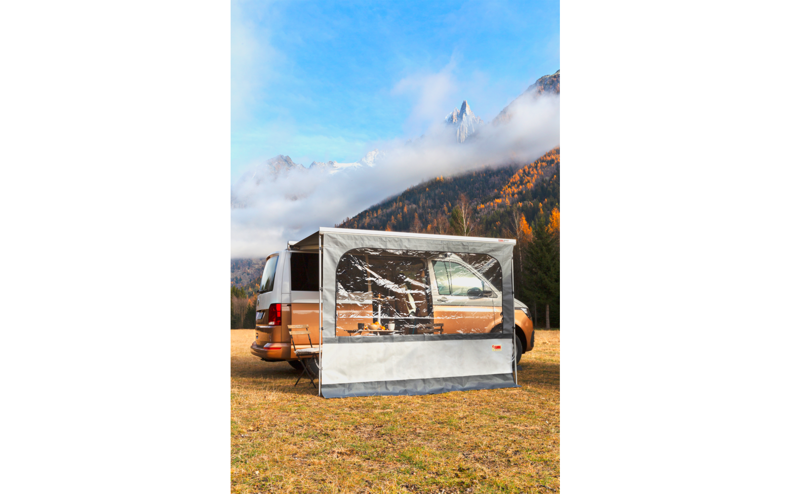 Weiß-brauner Campervan mit angebauter, durchsichtiger Markise; Tisch und Stühle sichtbar; auf Wiese vor herbstlichen Bäumen und nebelverhangenen Bergen
