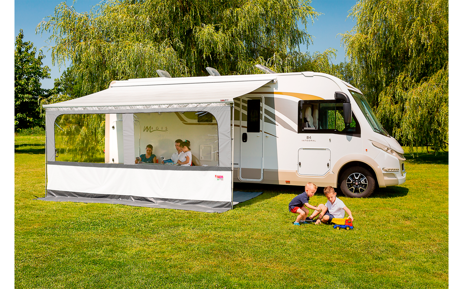 Wohnmobil auf einer Wiese mit ausgefahrener Markise und seitlichem Vorzelt; zwei Personen sitzen an einem Tisch im Vorzelt; zwei Personen sitzen auf dem Gras vor dem Wohnmobil und spielen mit Spielzeugautos; Bäume und blauer Himmel im Hintergrund