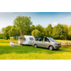 Silberner Van vor weißem Wohnwagen mit angebautem Vorzelt auf grüner Wiese neben einem Teich und einer Baumreihe unter blauem Himmel