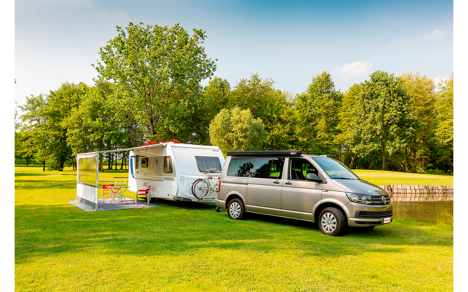 Silberner Van vor weißem Wohnwagen mit angebautem Vorzelt auf grüner Wiese neben einem Teich und einer Baumreihe unter blauem Himmel