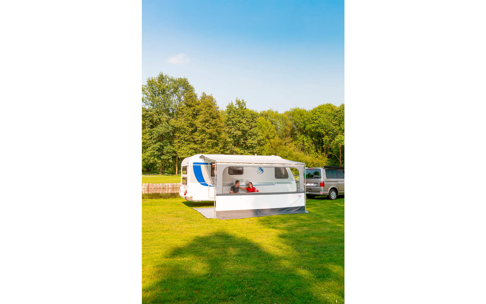 Wohnwagen auf grünem Rasen mit angebautem Vorzelt; zwei Personen sitzen im Vorzelt; ein Auto daneben; Bäume im Hintergrund; blauer Himmel
