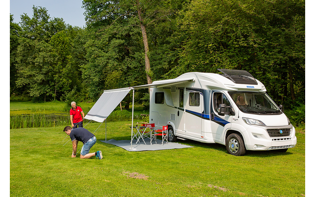 Weißes Wohnmobil auf einer Wiese mit ausgefahrener Markise, zwei Personen, die eine Bodenmatte auslegen; zwei rote Klappstühle, kleiner Tisch, Bäume und Teich im Hintergrund