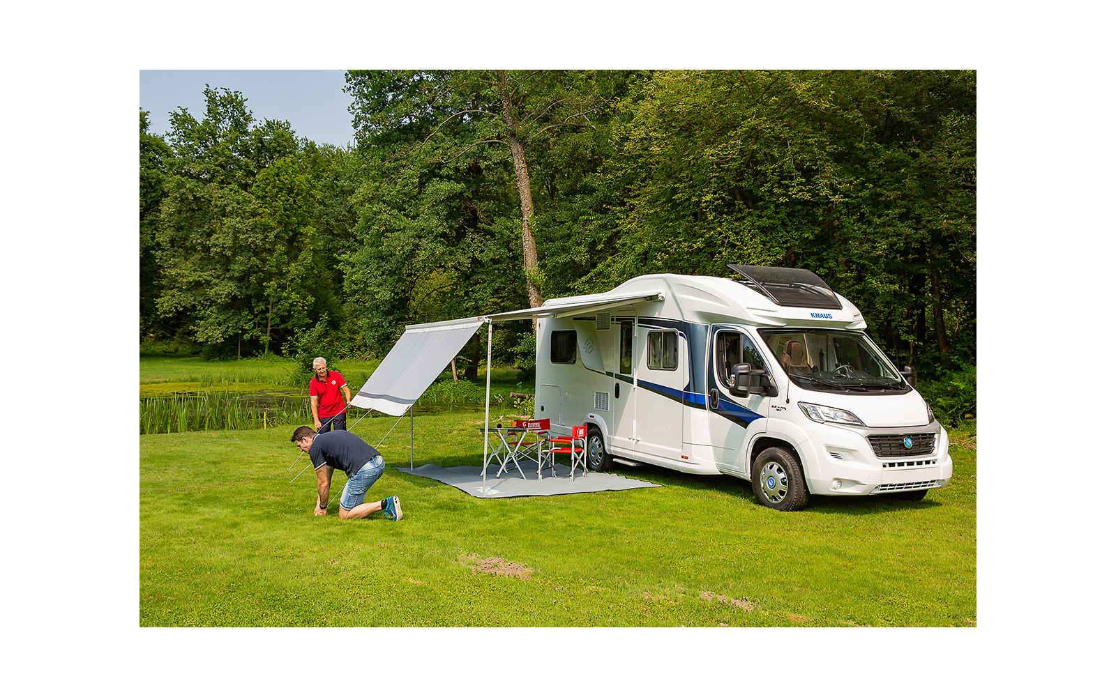 Witte camper op gras met uitgeklapte luifel, twee personen die een grondzeil uitrollen; twee rode klapstoelen, klein tafeltje, bomen en een vijver op de achtergrond