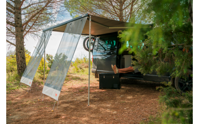 Dunkelgrauer Camper unter Bäumen mit ausgefahrener Markise, geöffnete Heckklappe, sichtbare nackte Beine, schwarzer Koffer und Fahrradreifen