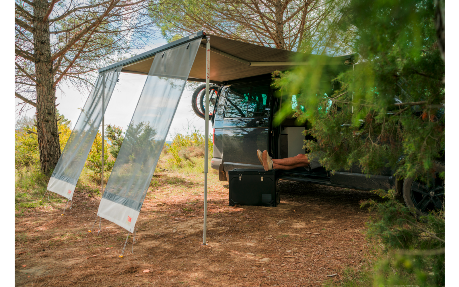 Dunkelgrauer Camper unter Bäumen mit ausgefahrener Markise, geöffnete Heckklappe, sichtbare nackte Beine, schwarzer Koffer und Fahrradreifen