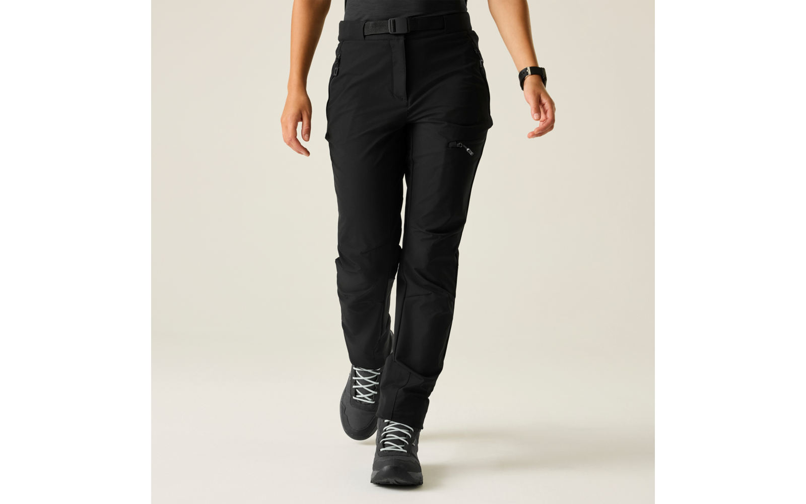 Onderlichaam van een persoon met zwarte lange broek met riem en ritszak op het bovenbeen, zwarte schoenen met lichte veters, handen en linkerpols met horloge zichtbaar, effen lichte achtergrond