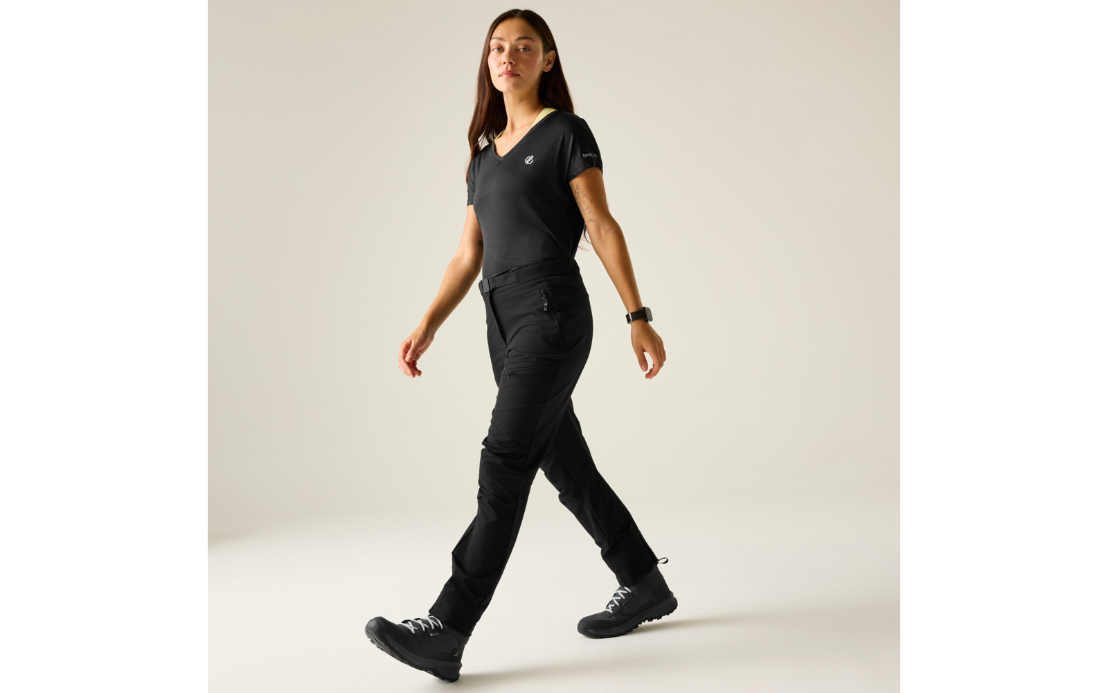 Volledige lichaamsfoto van een persoon die naar rechts loopt, met een zwart V-hals shirt, zwarte lange broek, zwarte laarzen, smartwatch om de linkerpols, effen beige achtergrond