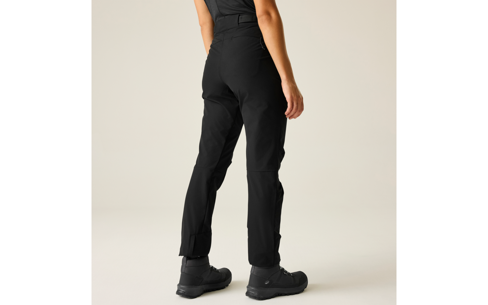 Achteraanzicht van een persoon van de taille naar beneden, zwarte outdoorbroek, zwarte enkellaarzen, lichte effen achtergrond