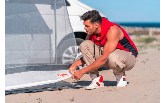 Ein Mann kniet im Sand neben einem weißen Wohnmobil und hält den Saum einer hellgrauen Zeltplane; Meer und blauer Himmel im Hintergrund